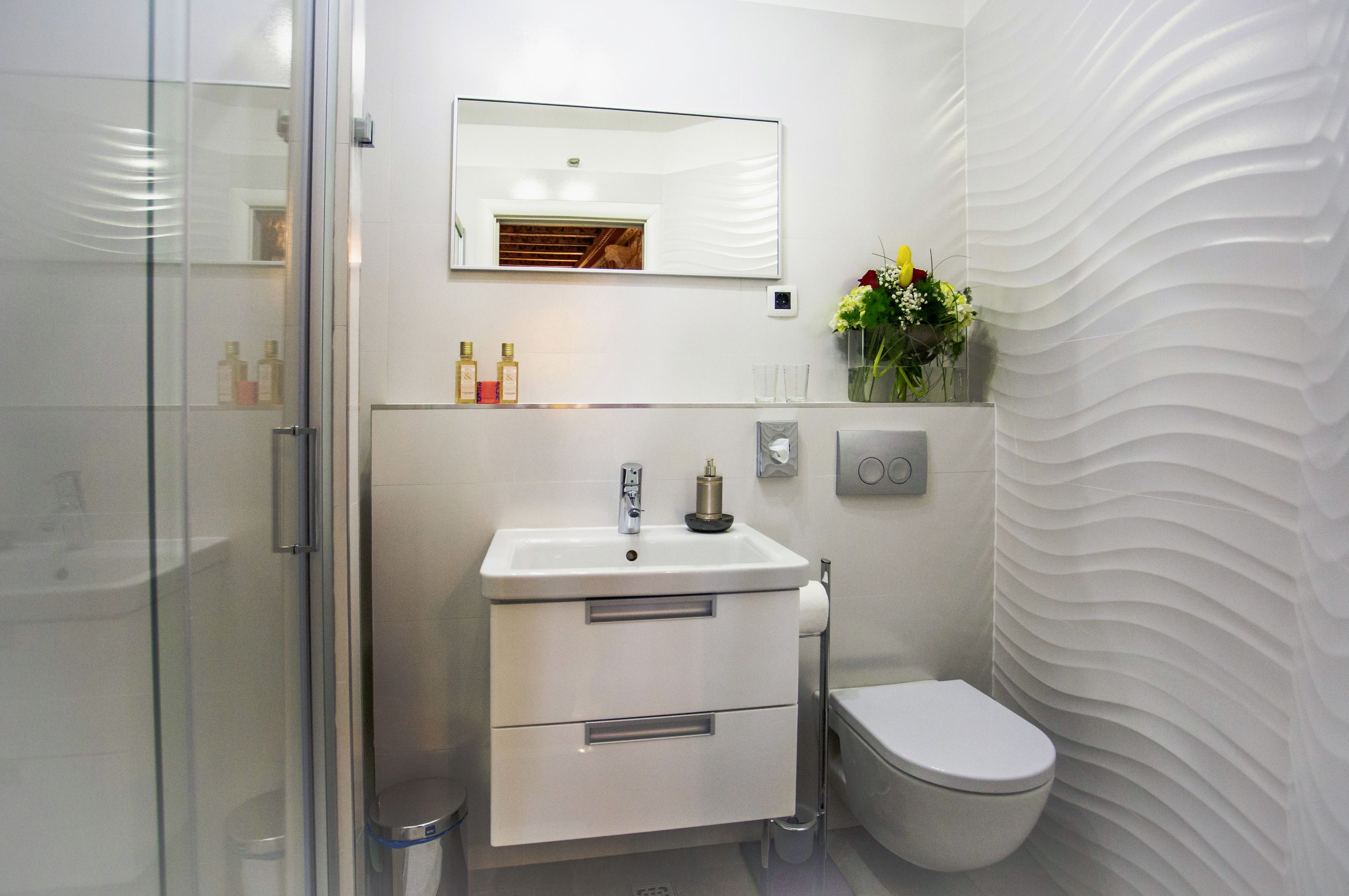 Une salle de bain moderne avec une douche à parois de verre, un lavabo blanc avec un miroir au-dessus, des toilettes et une étagère murale où sont posés des articles de toilette et un vase de fleurs.