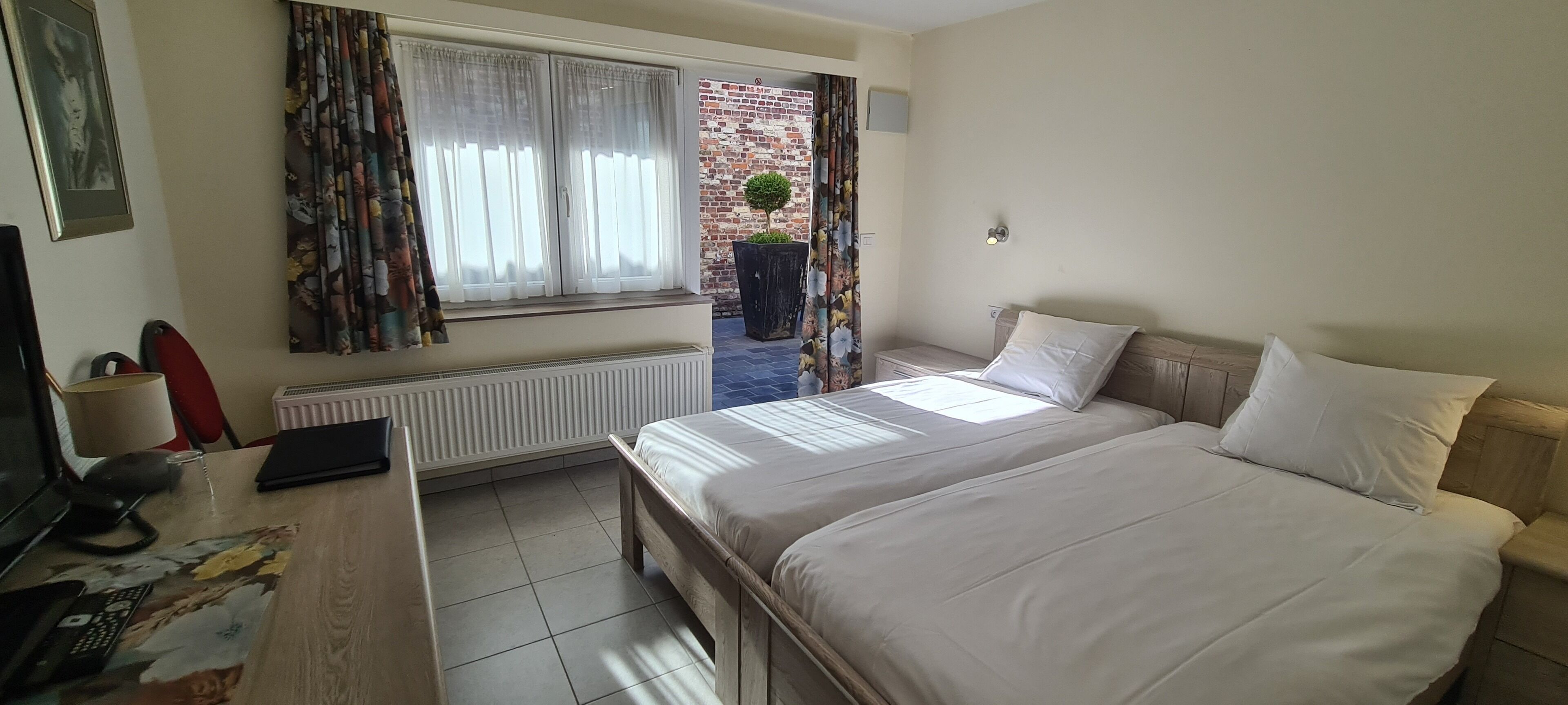 Une chambre d’hôtel avec un lit, un bureau avec un ordinateur, un radiateur et une fenêtre avec des rideaux.
