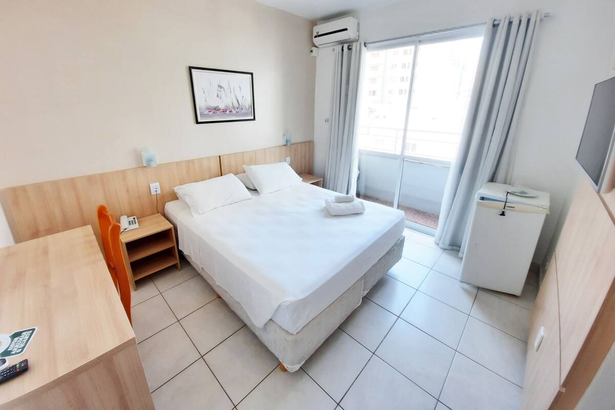 A hotel room with a bed, a desk, a chair, a TV, an air conditioning unit, and a window with curtains.