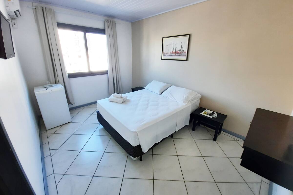 A hotel room with a bed, a nightstand, a small table, a heater, a window with curtains, and a wall-mounted picture.