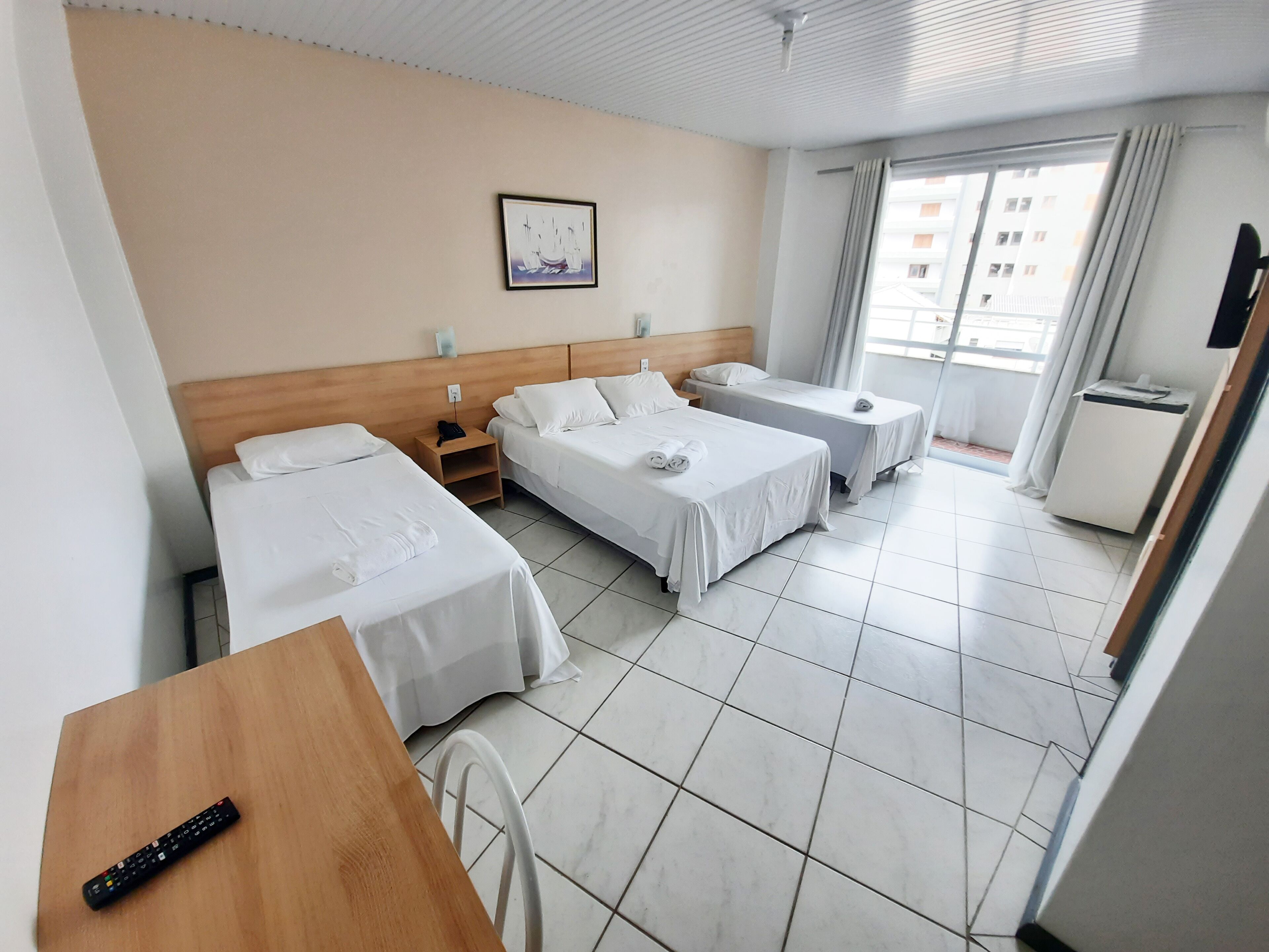 Habitación de hotel con tres camas, una mesa de madera, una silla, un televisor y una ventana con cortinas.