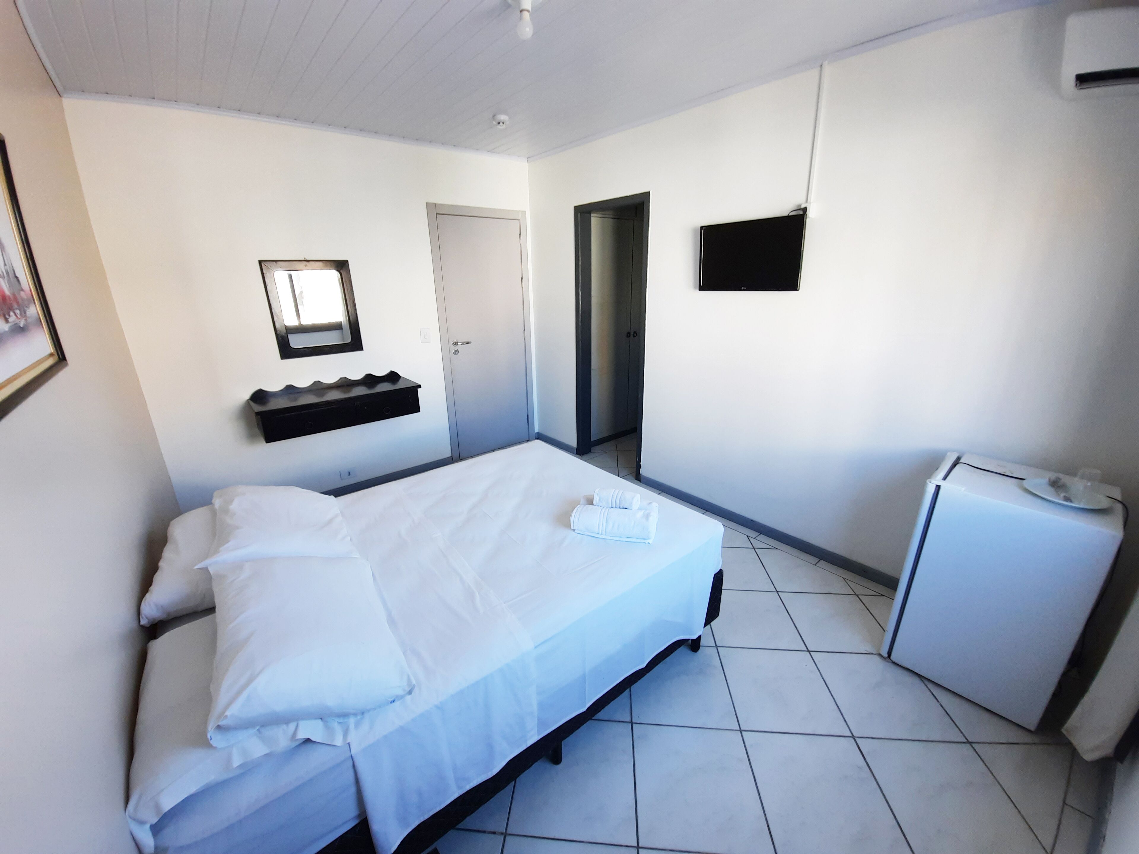 Un dormitorio con cama, un escritorio pequeño, un espejo, una puerta y un televisor en la pared.