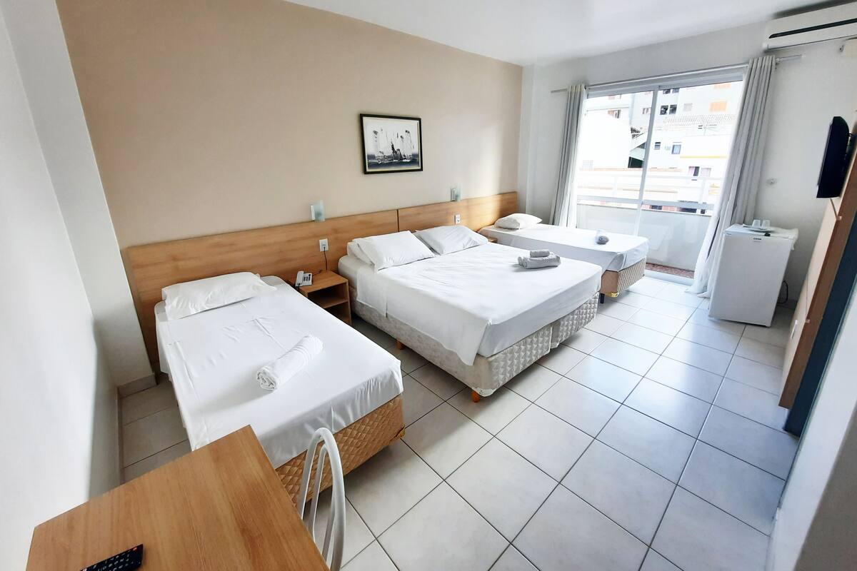 A hotel room with two beds, a desk, a chair, a small table, a refrigerator, and a window with curtains.