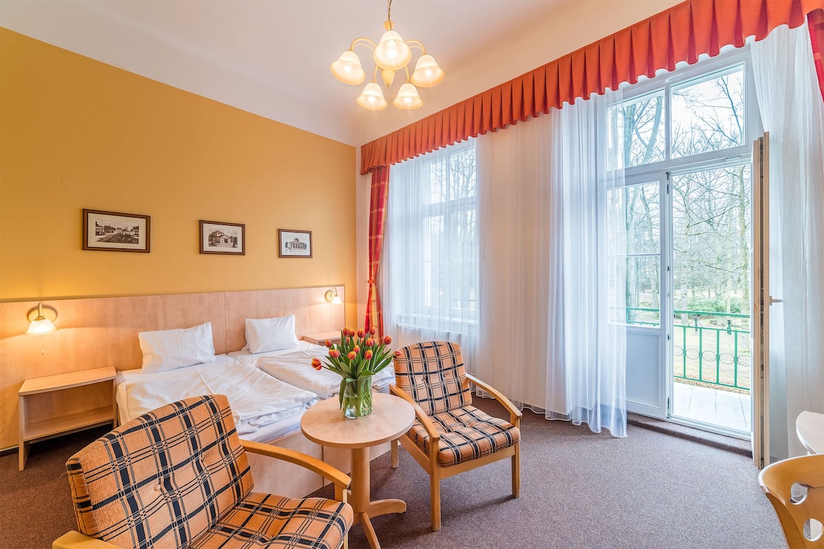 A hotel room with a bed, two chairs, a small table with a vase of flowers, and a window with curtains.