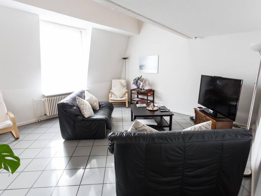 Una sala de estar moderna con un sofá de cuero negro, un sillón blanco, una mesa de centro de madera y un televisor de pantalla plana.