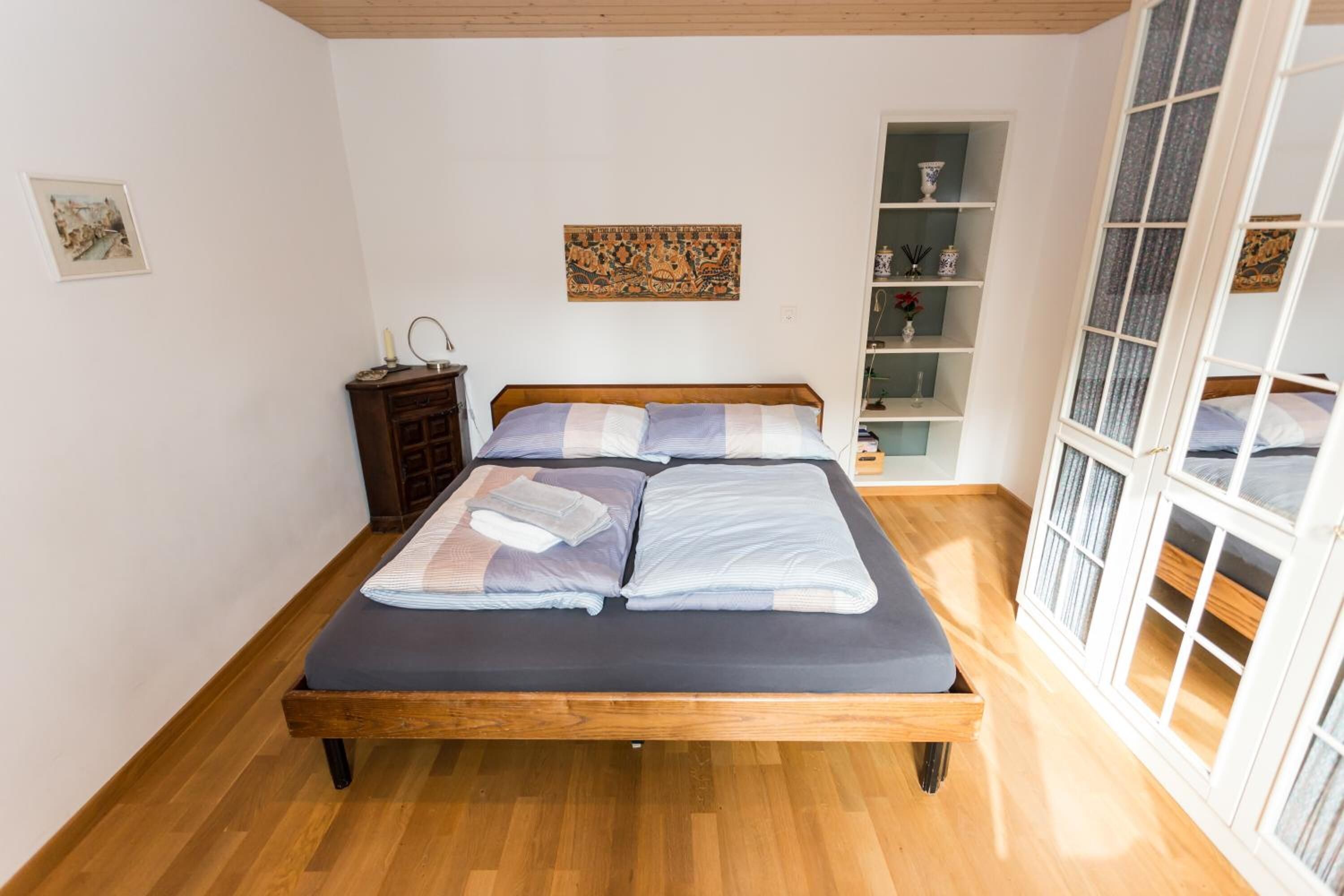 Un dormitorio con cama de madera, mesita de noche, estante con adornos y ventana con cortinas.