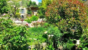 A lush garden with a variety of plants and flowers, a stone pathway, and a building with a sloped roof in the background. - Les Rosées (Mougins)