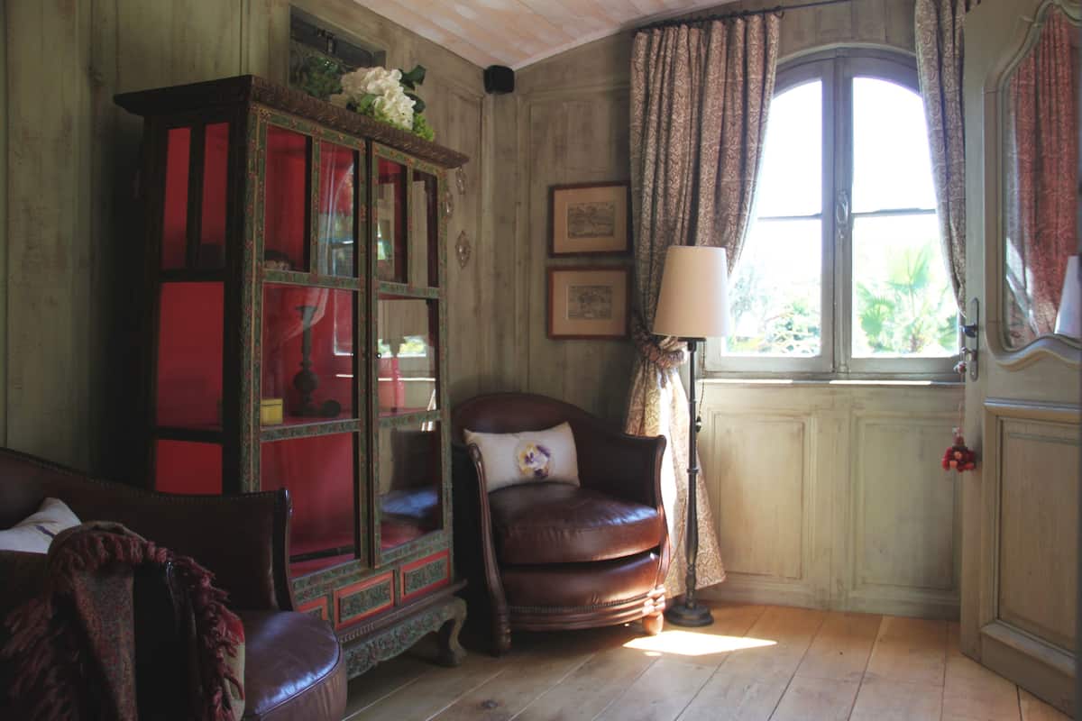 A room with a wooden floor, a leather armchair, a wooden cabinet with glass doors, a standing lamp, and a window with curtains.