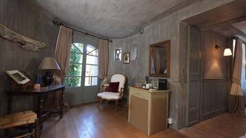 Ein Zimmer mit Holzboden, Schreibtisch mit Lampe, Stuhl, kleines Schränkchen und einem Fenster mit Vorhängen.