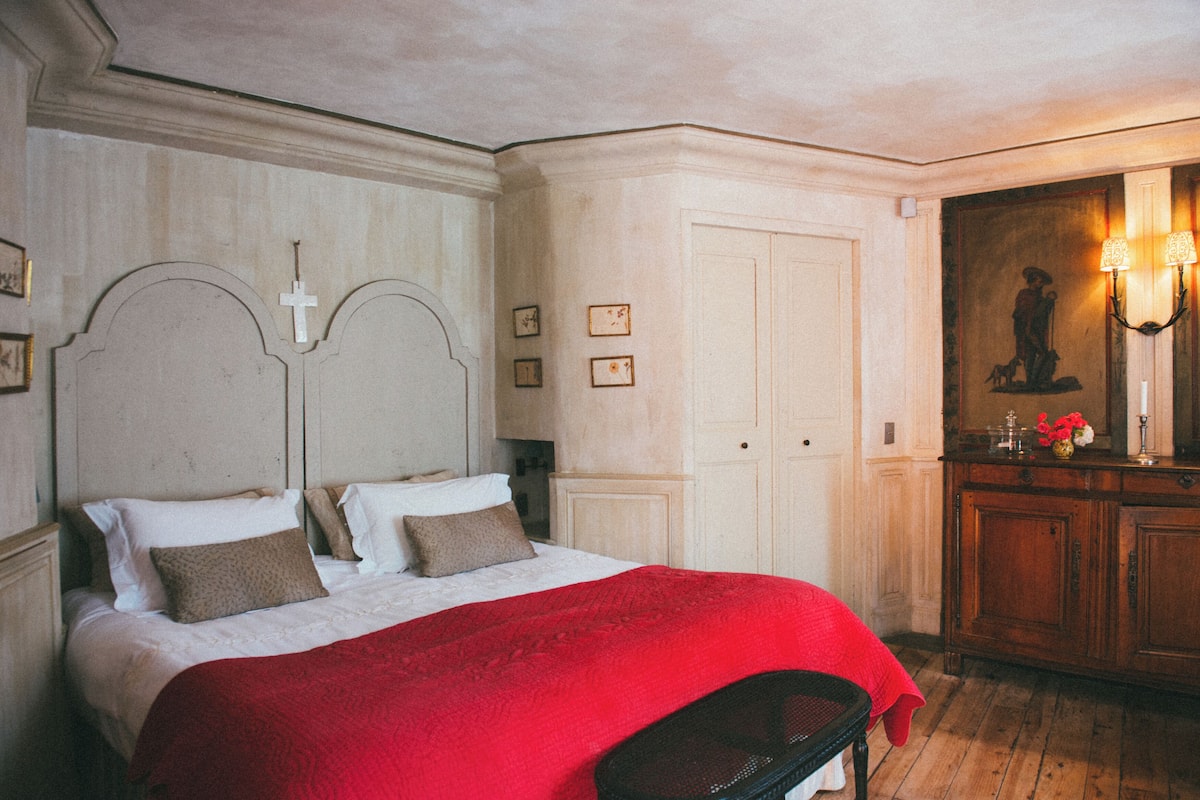 A bedroom with a bed, a red blanket, a wooden bench, a wall-mounted cross, and a painting.