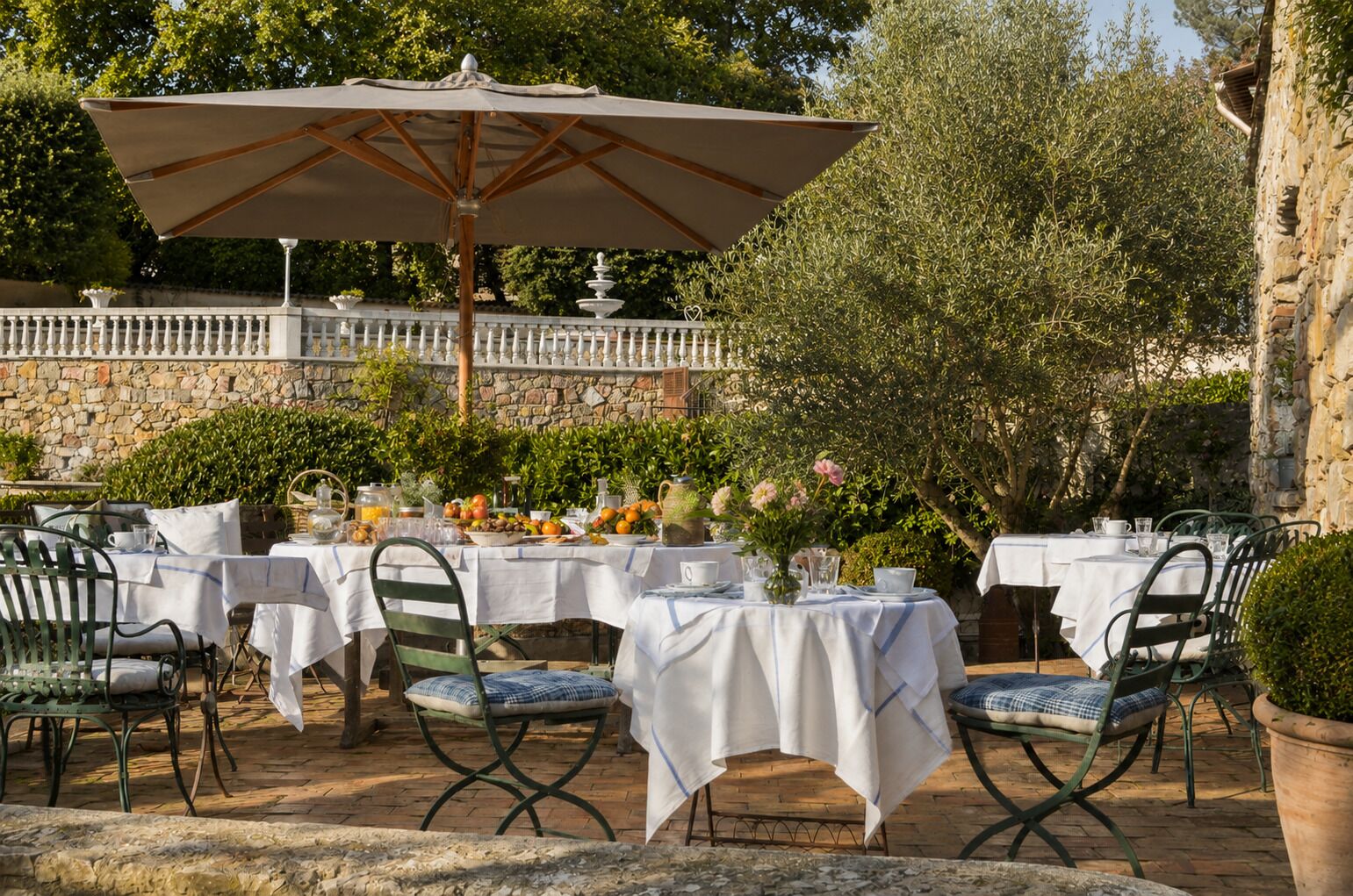 An outdoor dining area with tables and chairs, a large umbrella, and a stone wall in the background.