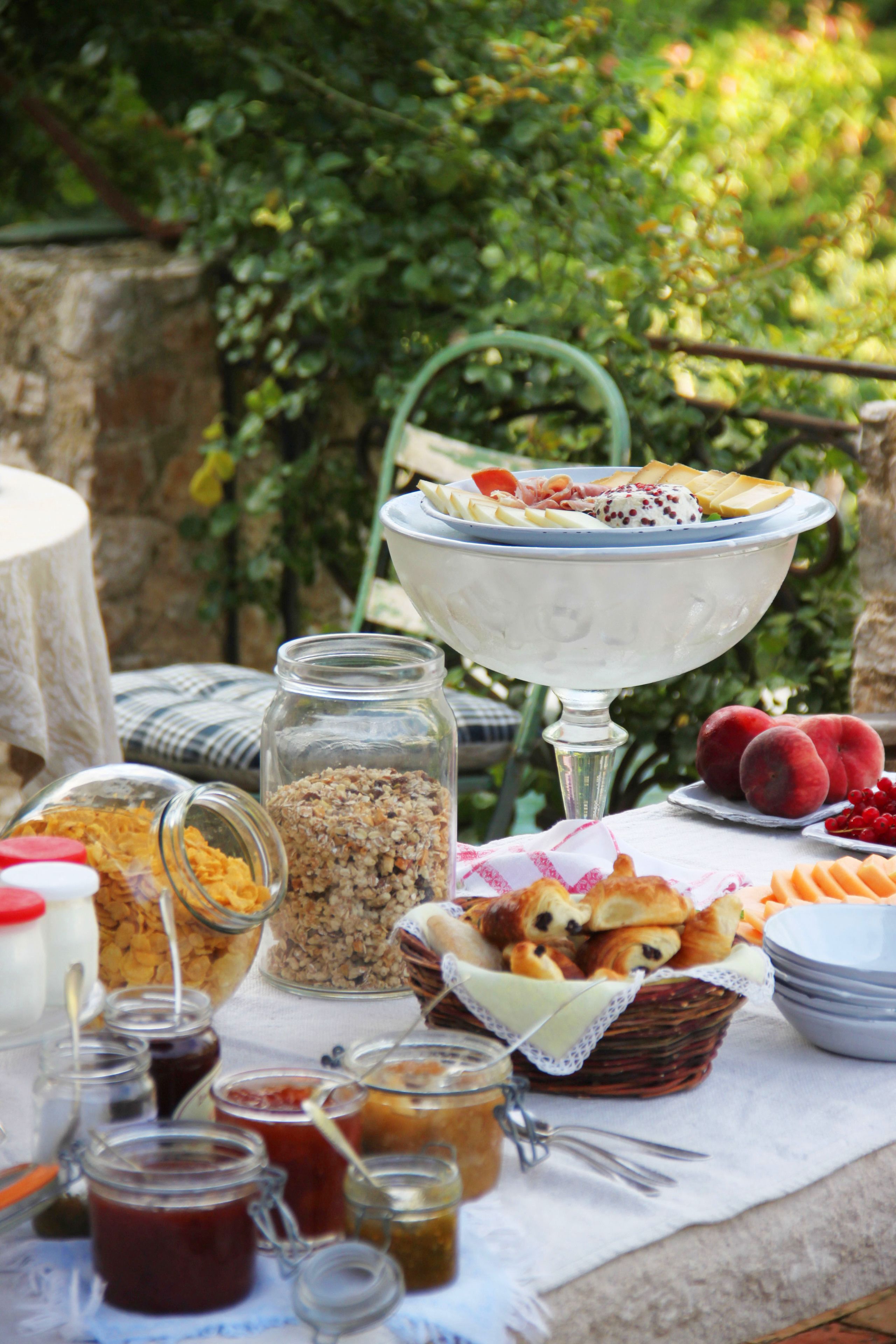 Una mesa al aire libre con diversas opciones de comida, incluyendo un cuenco de fruta, pasteles y frascos de conservas.