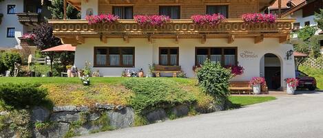 A traditional wooden chalet with balconies adorned with flowers, a well-maintained garden, and a clear view of the surrounding forest.