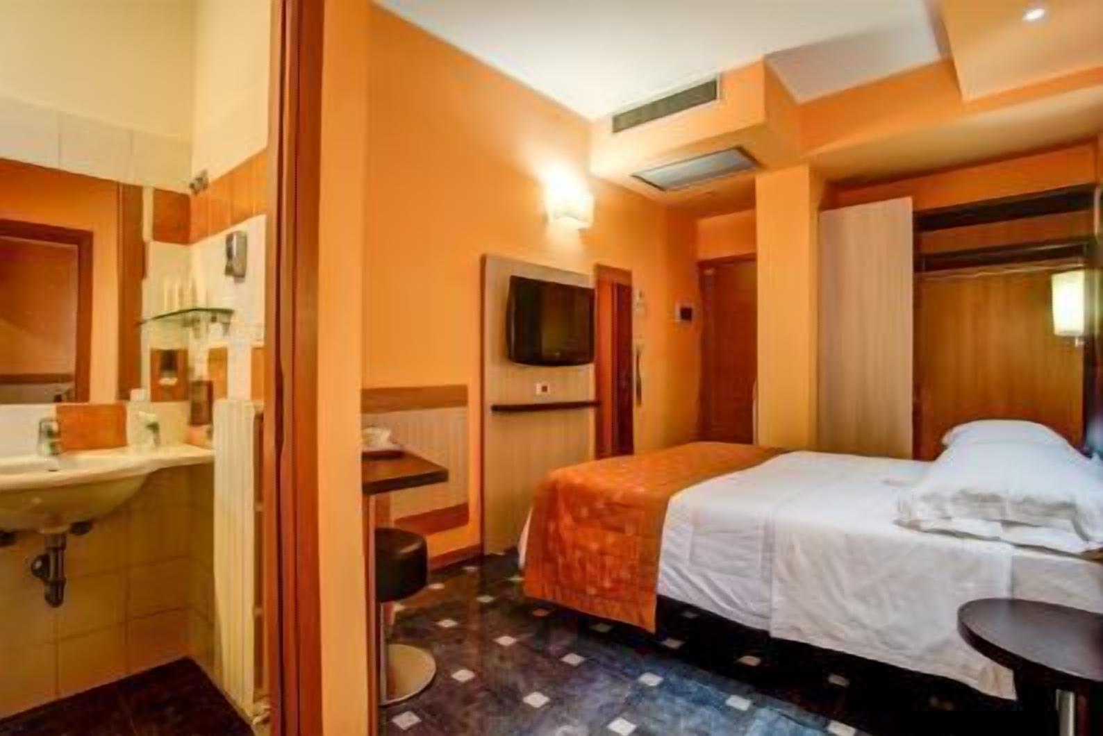 A hotel room with a bed, a small table, a chair, a TV, and a bathroom area.