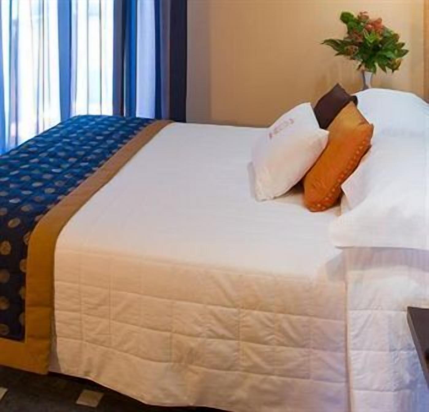 A neatly made bed with a white duvet, a blue patterned bedspread, and two pillows, one white and one orange.