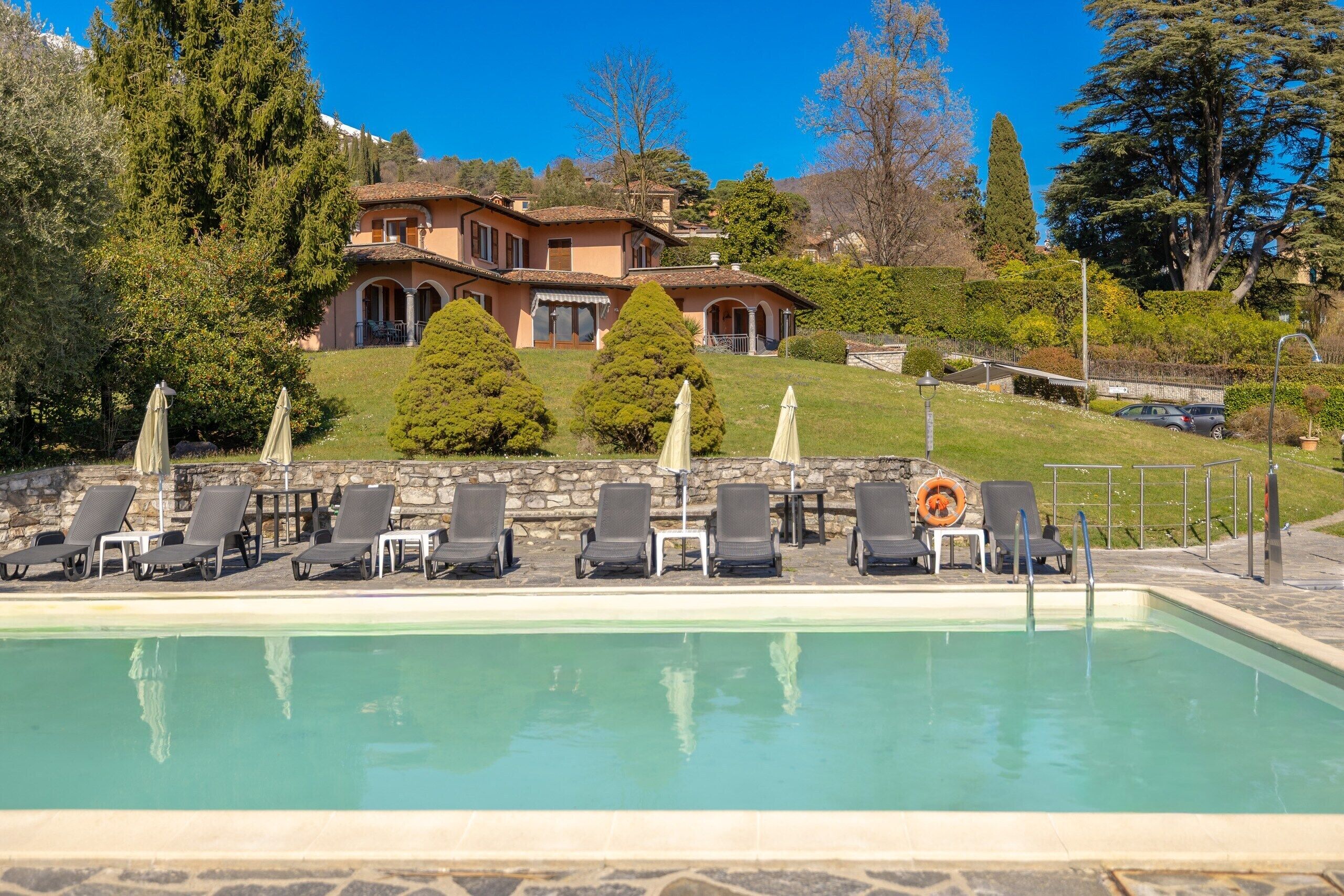 A pool with lounge chairs and umbrellas, a stone wall, and a building with a tiled roof in the background.