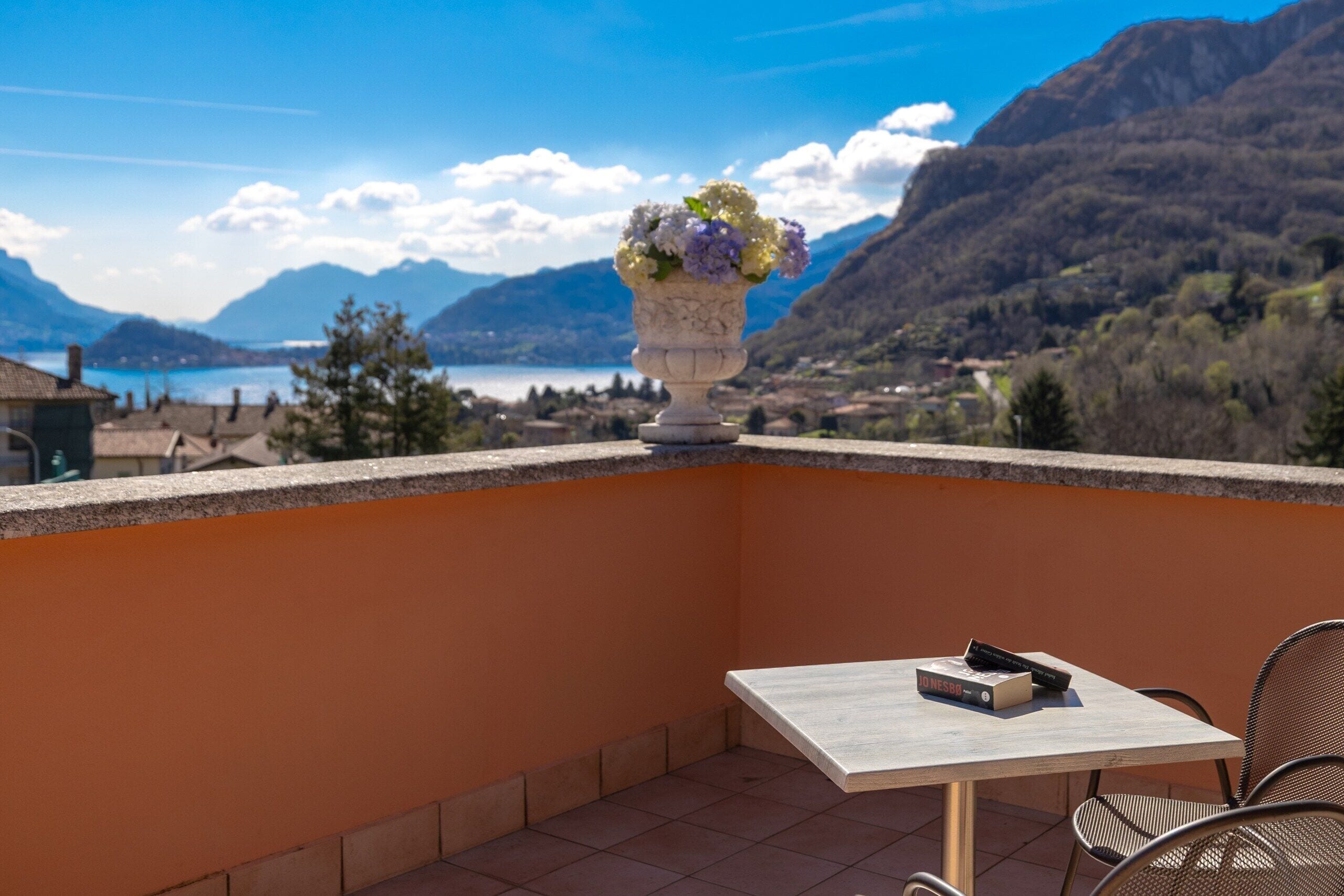 A balcony with a table, chairs, a vase of flowers, and a view of mountains and a lake.