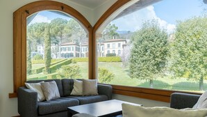 Quarto com sofá cinzento, uma mesa pequena e duas janelas amplas com vista para um jardim e edifícios.