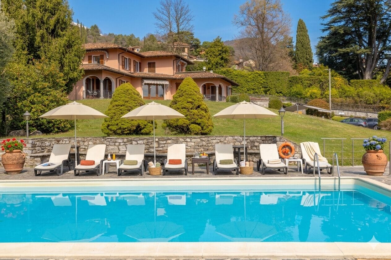 A pool with lounge chairs and umbrellas, a stone wall, and a large house in the background.