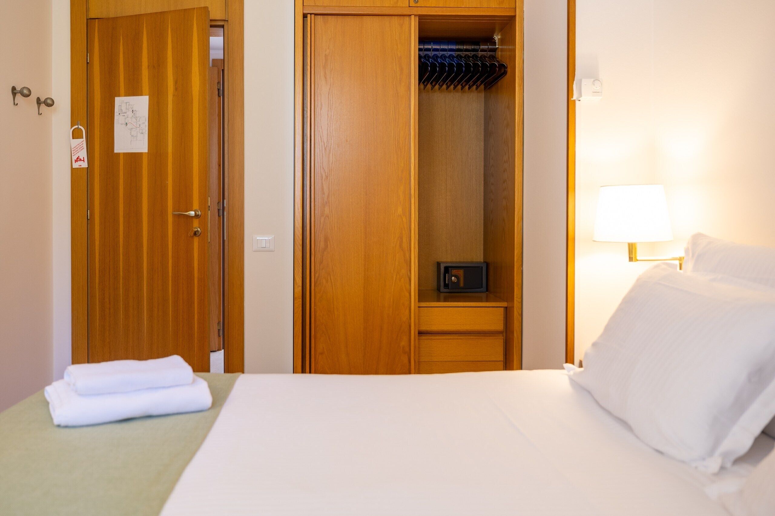 A hotel room with a bed, a wooden wardrobe, a bedside table with a lamp, and a wall-mounted clock.