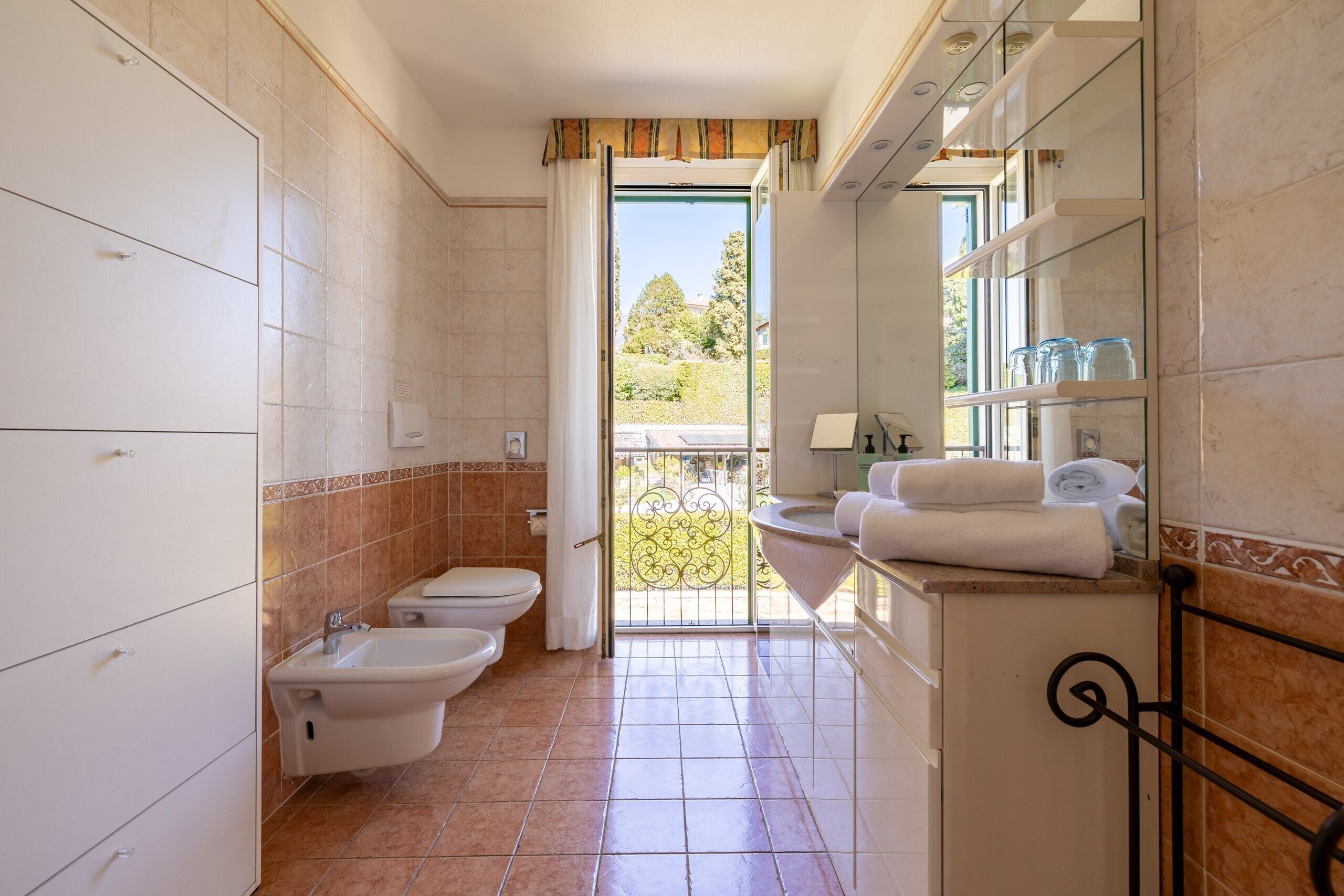 A bathroom with a toilet, bidet, and sink, a balcony with a view, and a tiled floor.