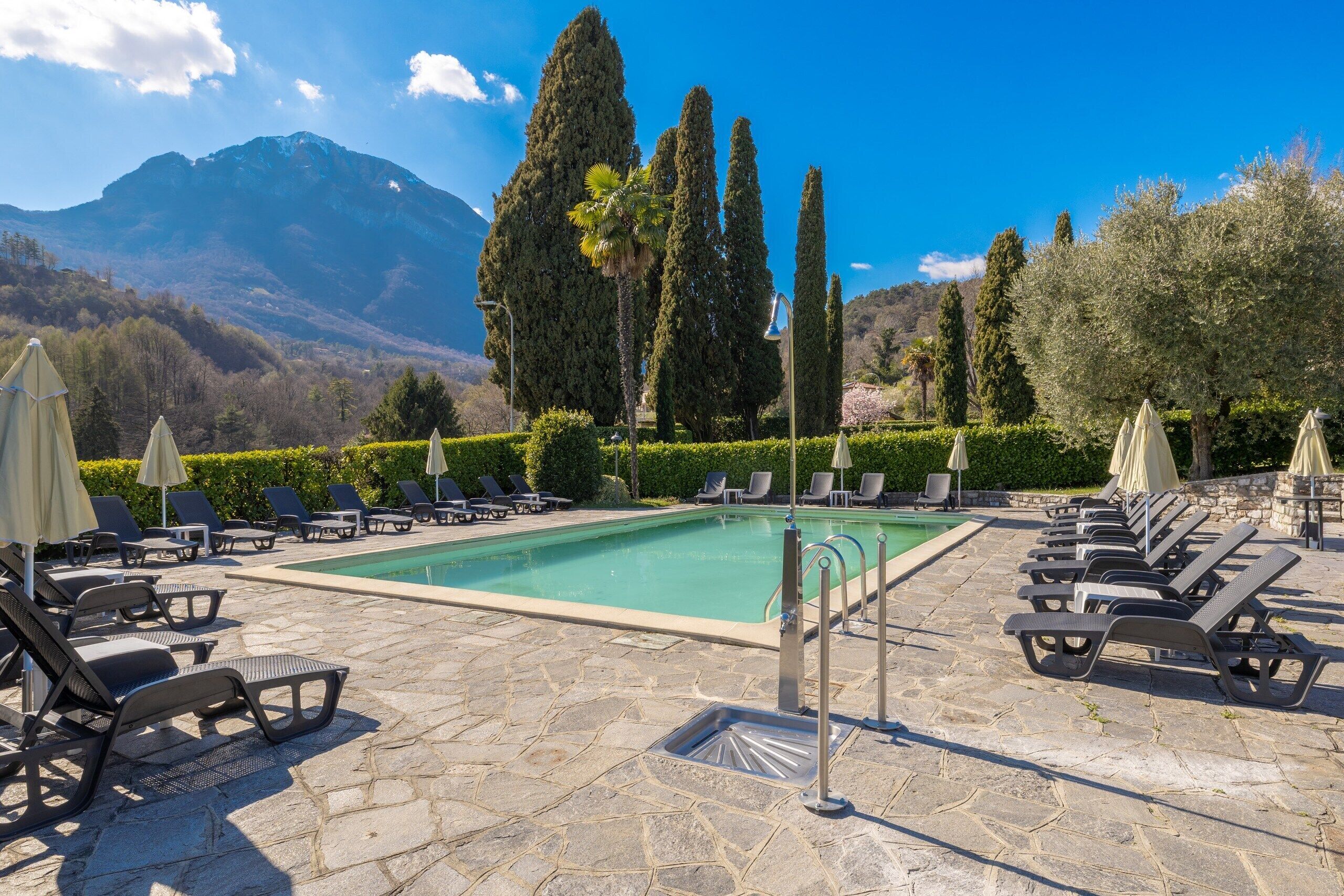 Un área de piscina con sillones de descanso y sombrillas, rodeada de setos bien cuidados y árboles altos, con montañas al fondo.