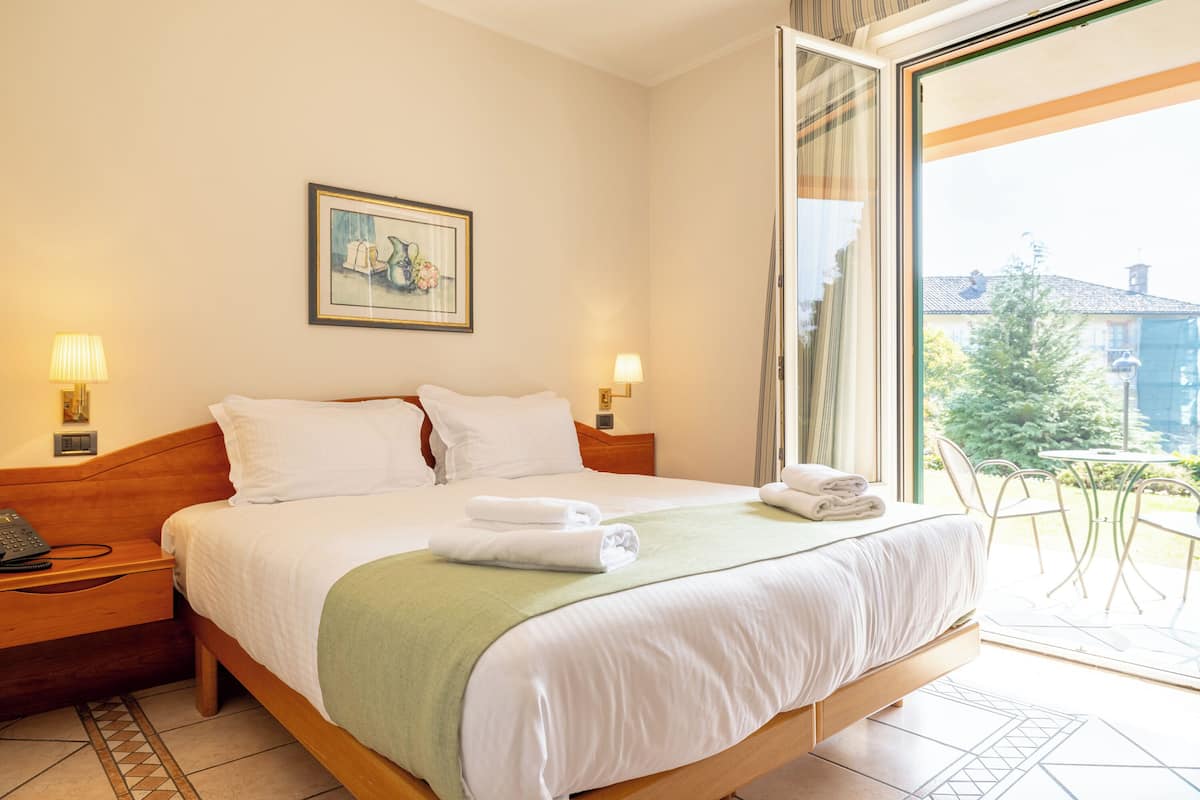 A hotel room with a large bed, two bedside tables with lamps, a green throw blanket, and a balcony with a view of buildings and trees.