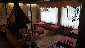 A dimly lit room with rustic brick walls, wooden tables, and chairs. A chandelier hangs from the ceiling, and there is a large window with sheer curtains.