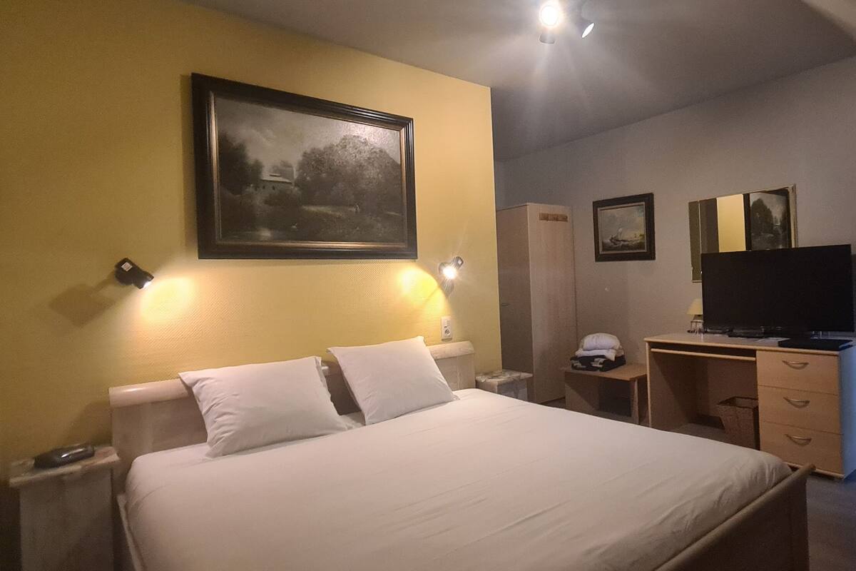 Een hotelkamer met een bed, nachtkastjes, een bureau met stoel, een televisie en beelden in lijsten aan de muur.