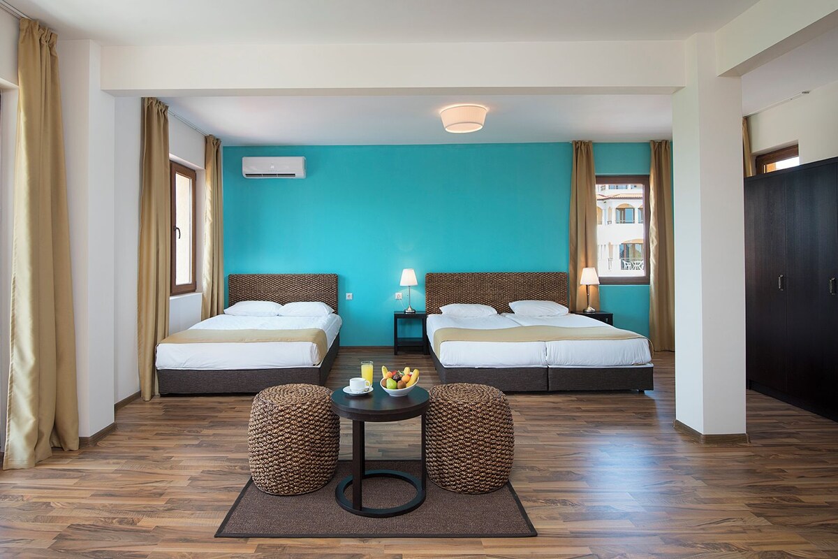 A hotel room with two beds, a small table with fruit, a view of the outdoors, and a window with curtains.