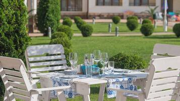 Una mesa al aire libre con sillas blancas, mantel azul y blanco, y vasos de vidrio transparente.