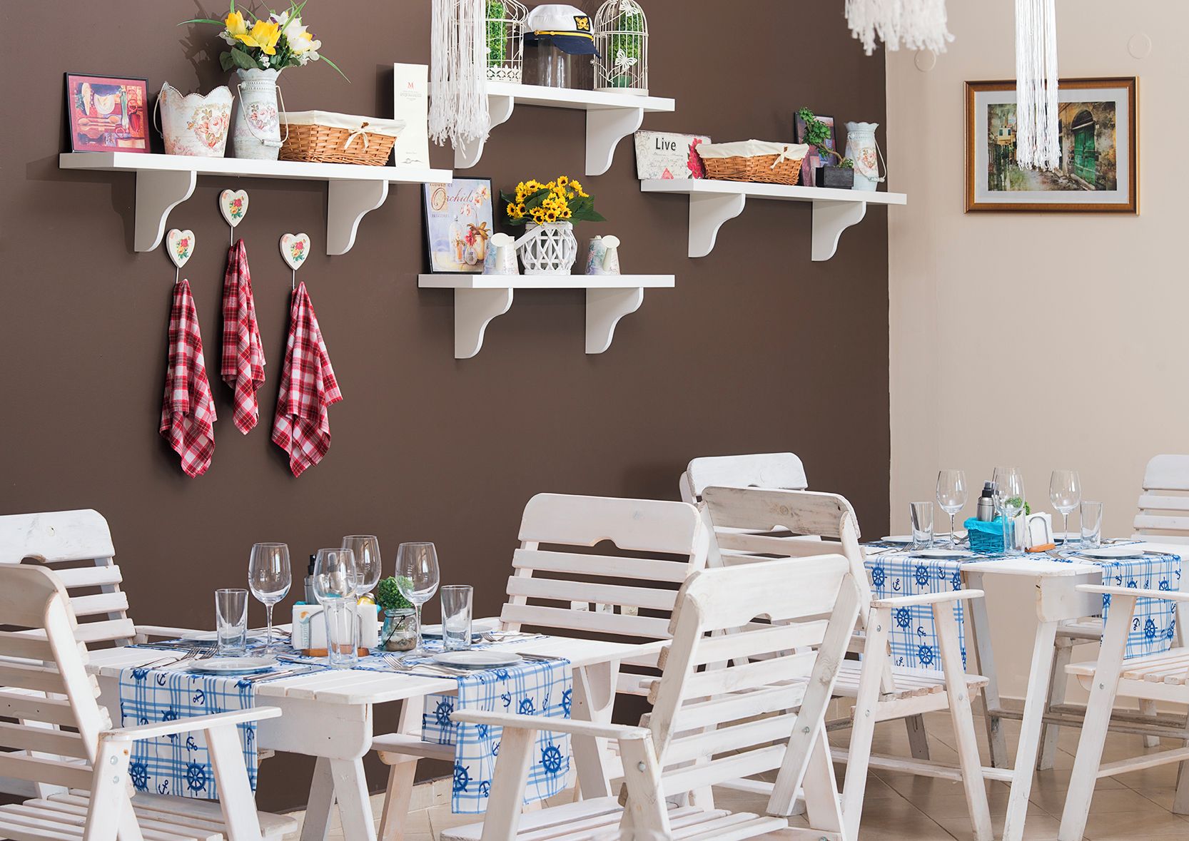 Un comedor con sillas y mesas de madera blanca, algunas con manteles a cuadros azules y blancos. Hay estanterías blancas con varios objetos, incluyendo cestas y adornos.
