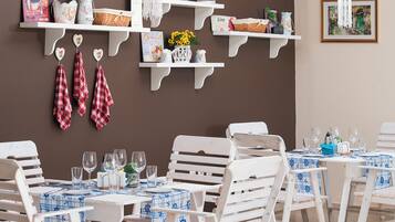 Un comedor con sillas y mesas de madera blanca, algunas con manteles a cuadros azules y blancos. Hay estanterías blancas con varios objetos, incluyendo cestas y adornos.