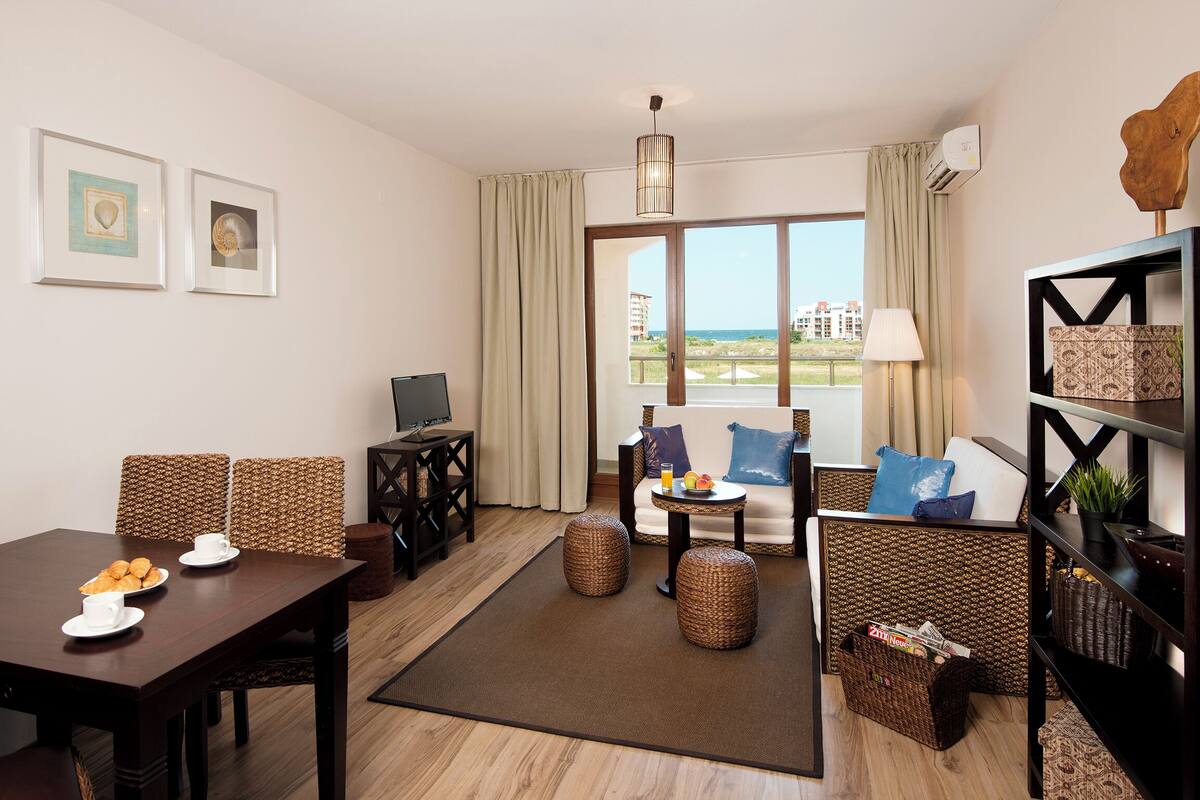 A modern living room with a dining area, a TV, wicker furniture, and a view of the sea.