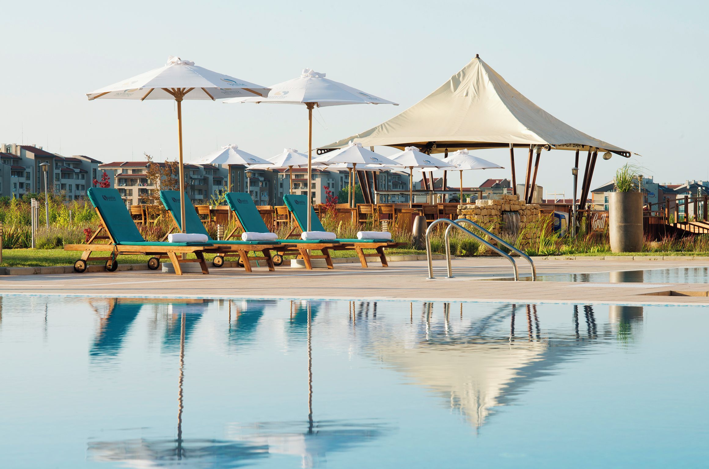A poolside area with lounge chairs and umbrellas, a clear pool, and a backdrop of buildings.