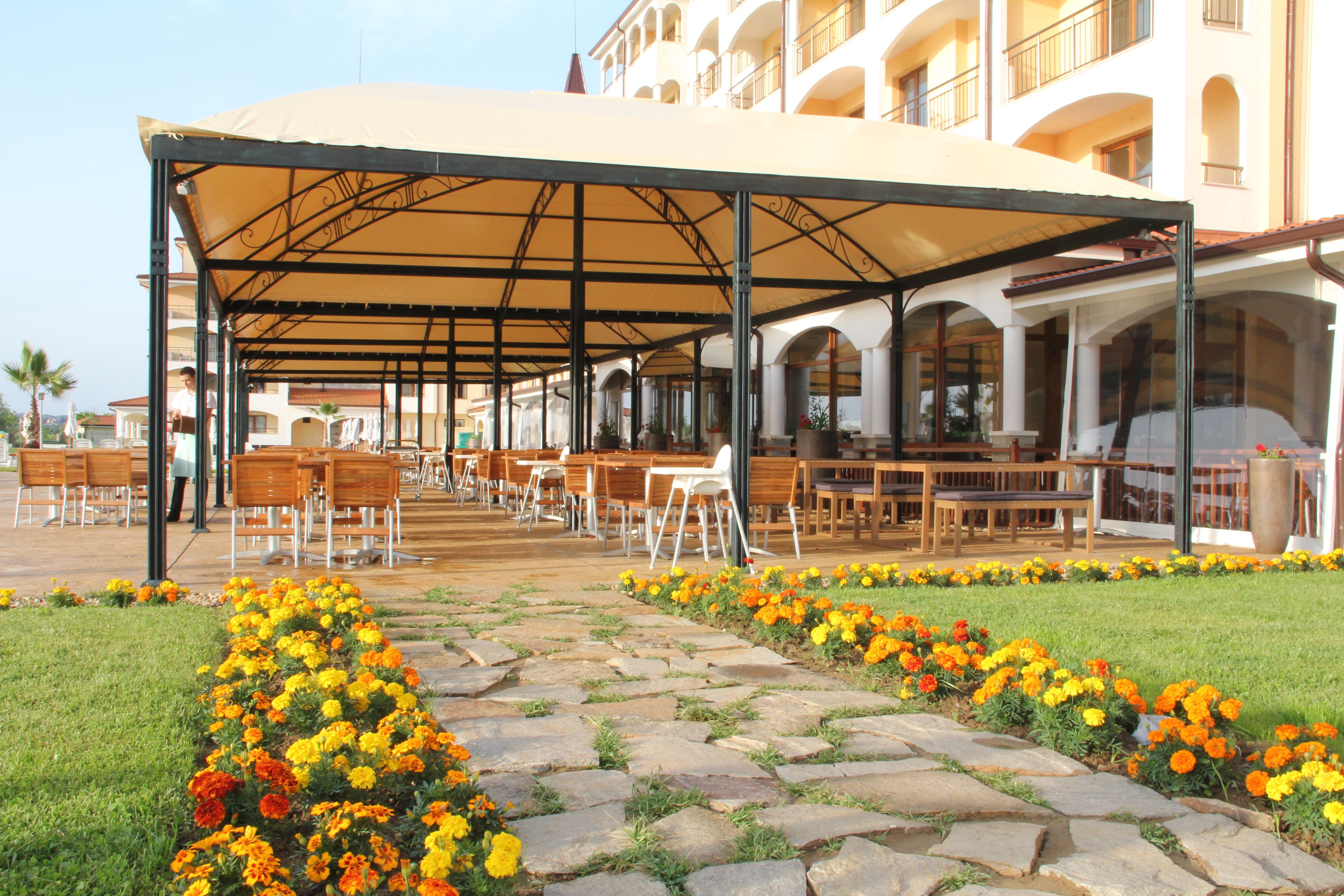 An outdoor seating area with tables and chairs under a covered structure, surrounded by flower beds.