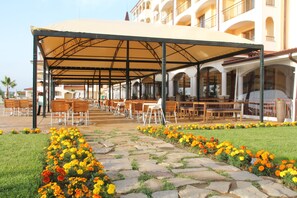 An outdoor seating area with tables and chairs under a covered structure, surrounded by flower beds. - Sunrise All Suites Resort (Nessebar)