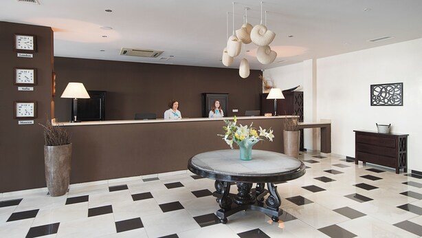 Ein Hotel-Empfangsbereich mit einer Rezeption, zwei Angestellten, einem runden Tisch mit Blumen und einem gemusterten Boden.