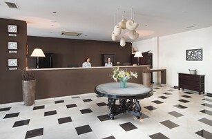 A hotel reception area with a check-in counter, two receptionists, a round table with a vase of flowers, and a patterned floor.