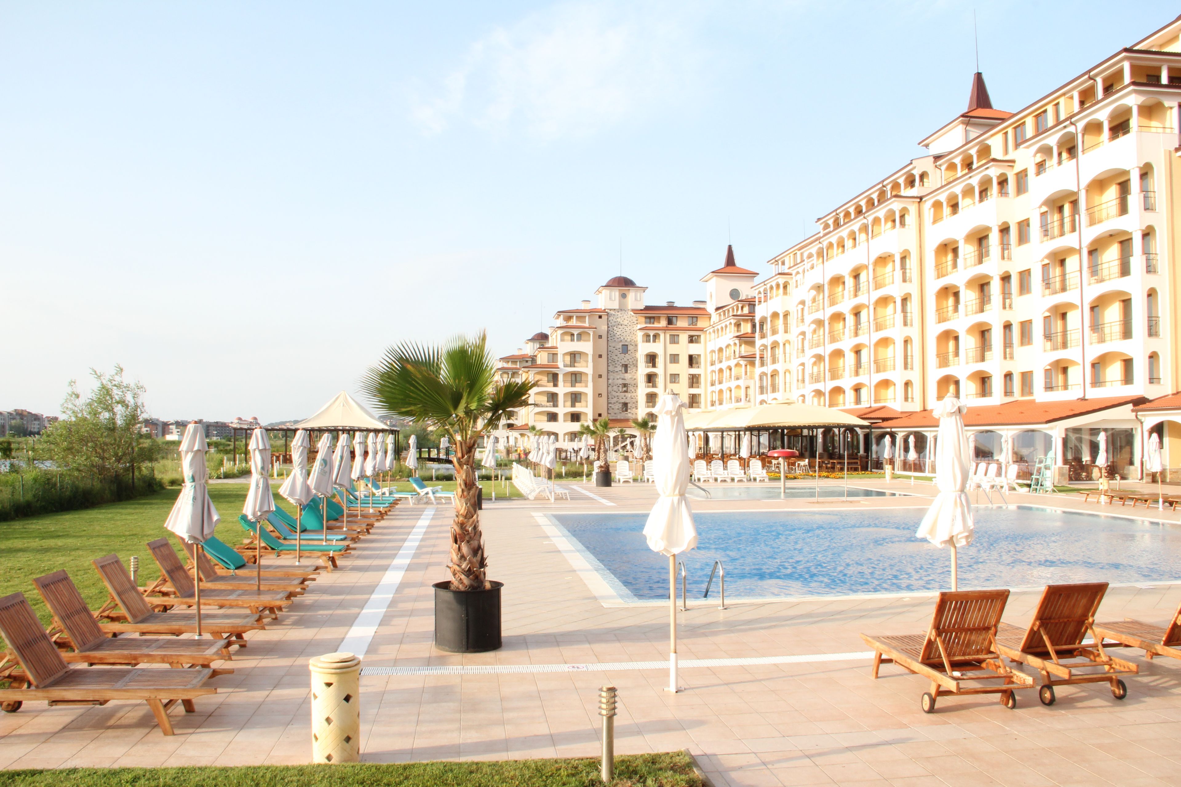 Zona de piscina hotelera con tumbonas y sombrillas, una palmera y un edificio grande con varios balcones.