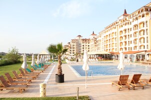 Zona de piscina hotelera con tumbonas y sombrillas, una palmera y un edificio grande con varios balcones.