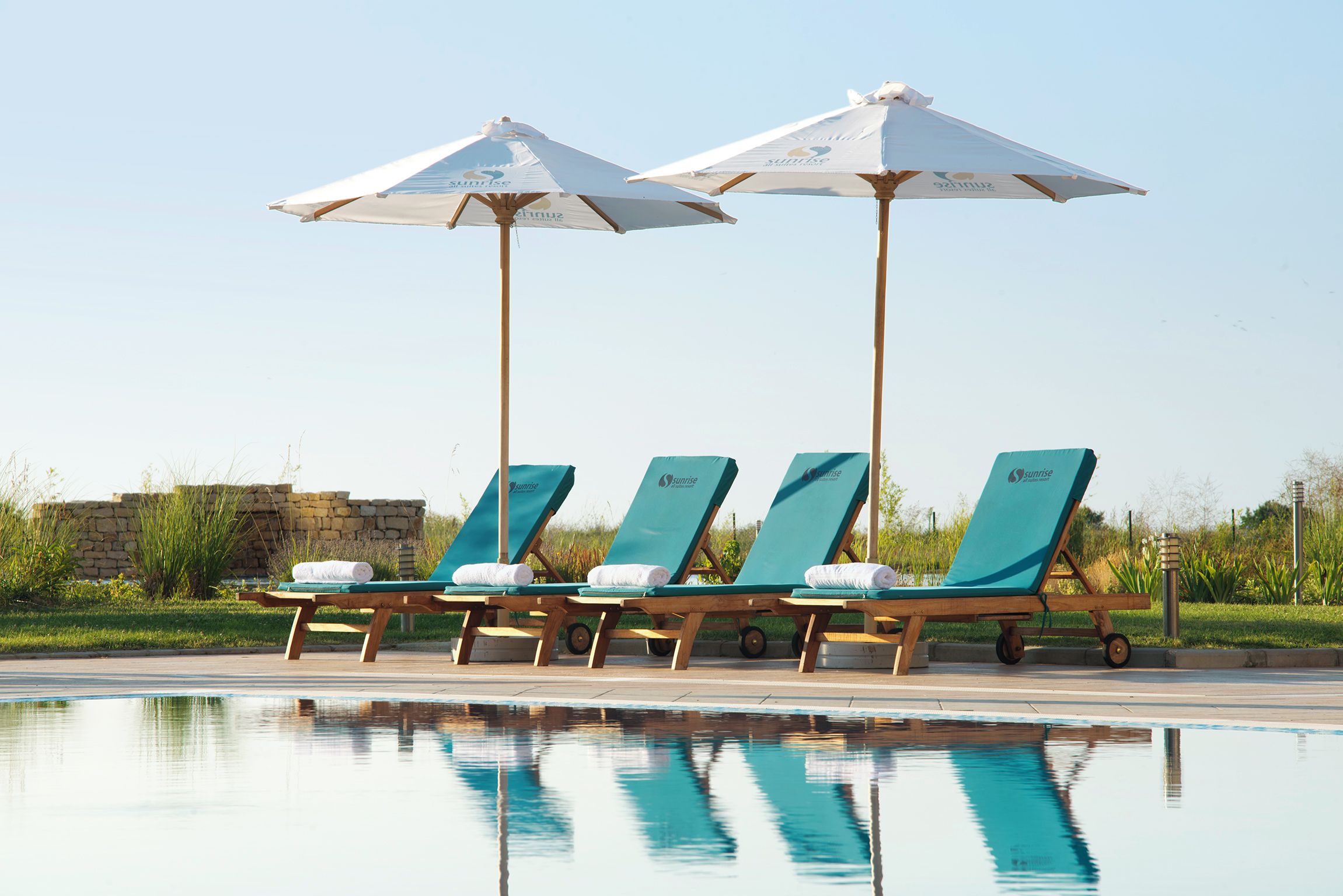 A poolside area with lounge chairs and umbrellas, a stone wall, and a clear sky.