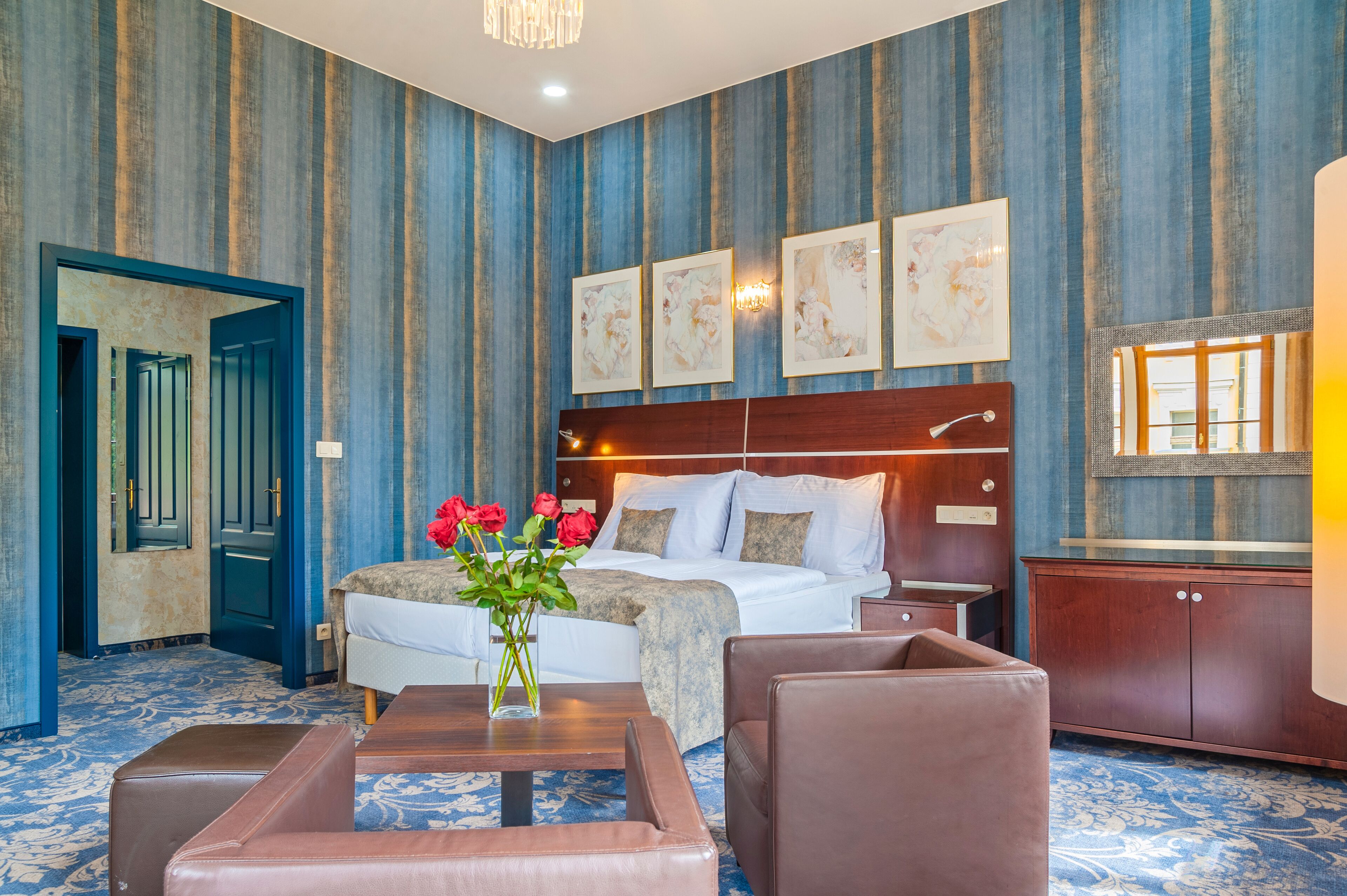 A hotel room with a large bed, a wooden headboard, a nightstand, a desk, a chair, a vase with flowers, and a large mirror.