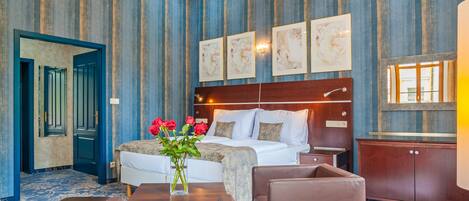 A hotel room with a large bed, a wooden headboard, a nightstand, a desk, a chair, a vase with flowers, and a large mirror.