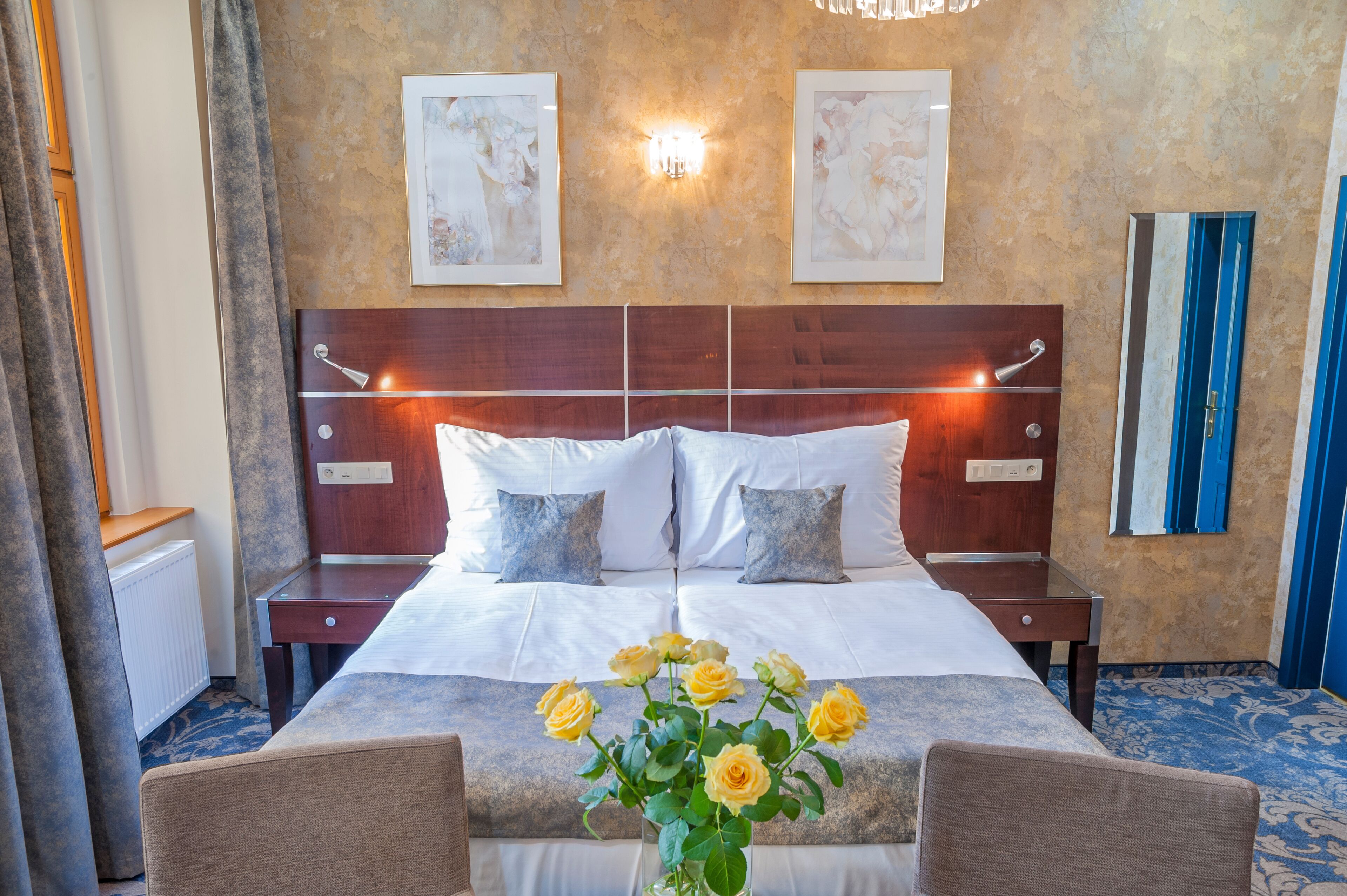A hotel room with a large bed, two bedside tables, a nightstand, a chair, a vase with flowers, and artwork on the walls.