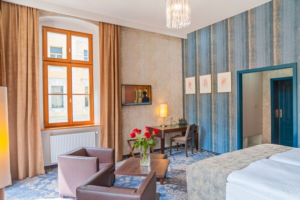 Hotelkamer met een bed, een bureau met een lamp, een stoel, een vaas met bloemen en een groot raam met gordijnen.