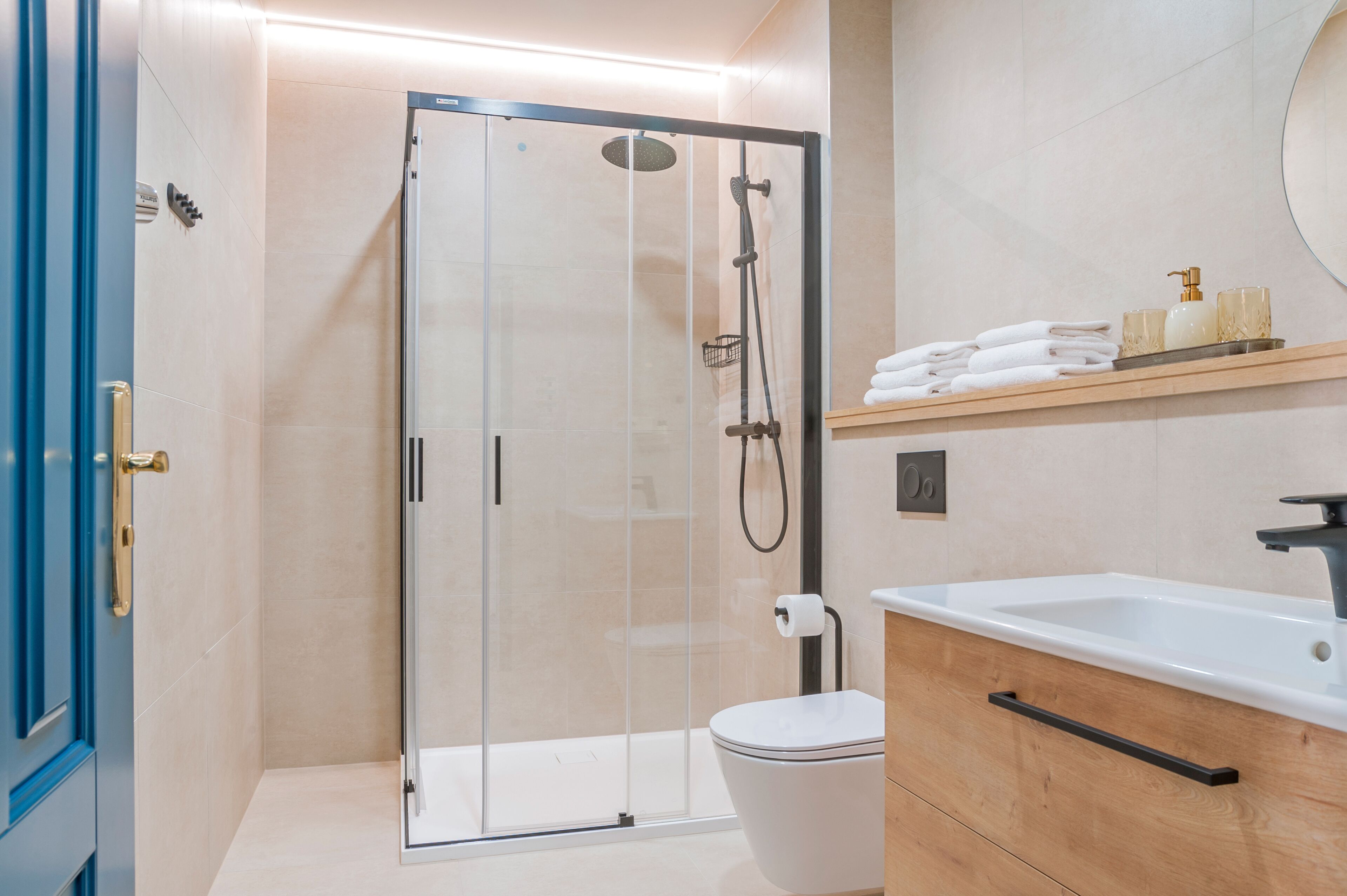 A modern bathroom with a glass-enclosed shower, a white toilet, a wooden vanity with a sink, and a shelf with towels and toiletries.
