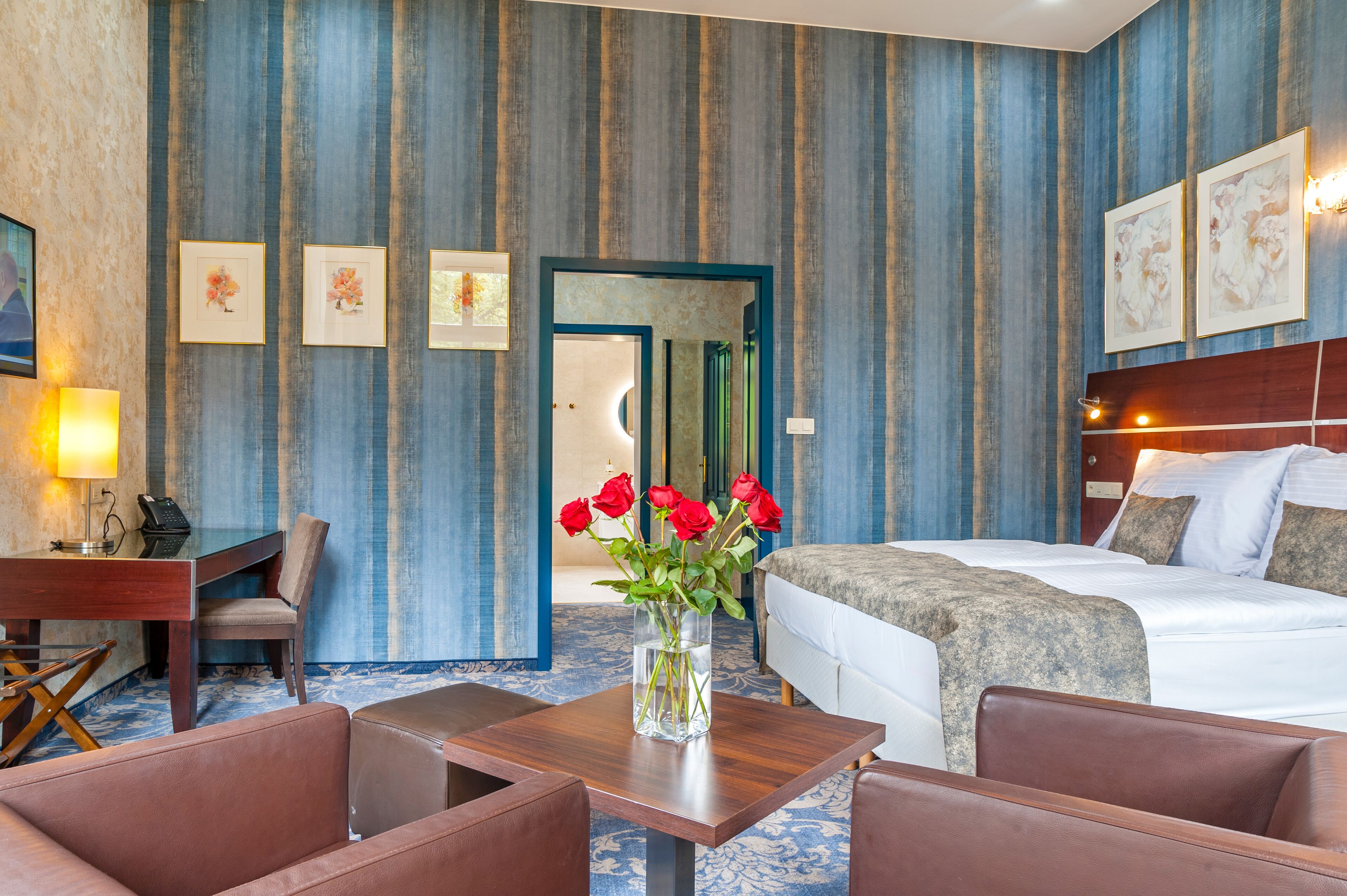 A hotel room with a bed, a desk, a chair, a table, and a vase with red flowers.