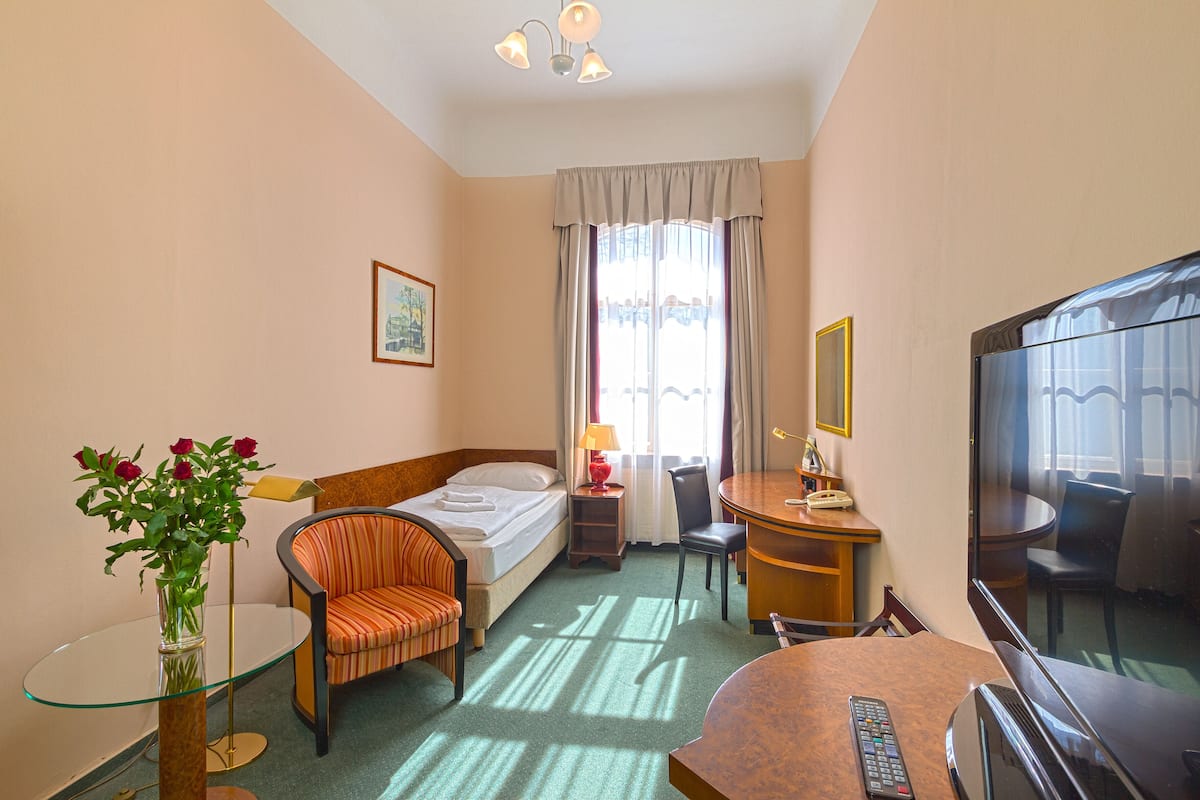 Ein Hotelzimmer mit einem Bett, einem Sessel, einem Schreibtisch, einem Fernseher, einem Glastisch, einer Lampe, einer Vase mit Blumen und einem Fenster mit Vorhängen.