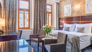 Une chambre d’hôtel avec un lit, un bureau et deux chaises. Il y a une fenêtre avec des rideaux, une lampe fixée au mur et un vase avec des fleurs sur le bureau.